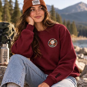 May include: A maroon crewneck sweatshirt featuring a circular patch on the chest with an illustration of an opossum and the text 002. The left sleeve has the text #002 printed in black. The sweatshirt is paired with light blue denim jeans.