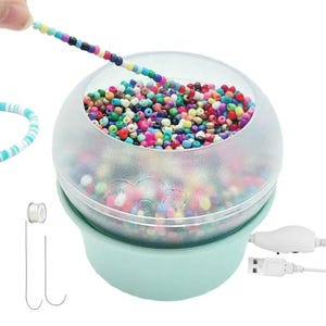 May include: An electric bead spinner with a light mint green base and a clear, rounded bowl filled with a variety of small, multi-colored plastic beads. A white USB power cable is attached, and two thin metal needles are included for threading.