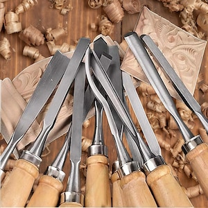 May include: A collection of wood carving chisels with polished metal blades and light-colored wooden handles, arranged in a fan shape. The tools feature various blade profiles, including straight, curved, and gouge shapes, resting on wood shavings.