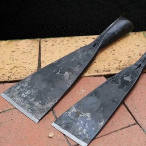 May include: Two hand-forged metal scrapers with dark, weathered surfaces and wide, flat blades. Each tool features a tapered, integrated handle that transitions into a broad, sharpened edge, showing signs of oxidation and a rustic, industrial finish.