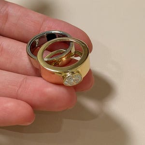 Puede incluir: Un anillo de tono dorado con una banda ancha y un engaste circular texturizado, colocado frente a un anillo de tono plateado liso. Ambos anillos presentan un acabado metálico pulido y un diseño simple y redondeado con una cara circular plana.