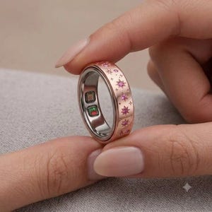 Peut inclure: Une bague connectée de couleur or rose avec une bande gravée de motifs en forme d'étoile, chacun orné d'une petite pierre précieuse rose. L'intérieur de la bague présente deux petits capteurs carrés, l'un avec une lumière verte et l'autre rouge.