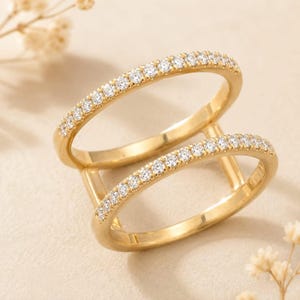 Puede incluir: Un anillo de oro de doble banda con dos aros paralelos unidos por barras verticales. Cada banda está engastada con una hilera de pequeñas piedras transparentes de corte redondo en la superficie superior, creando un brillo continuo sobre el metal.