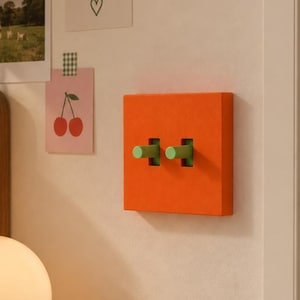 May include: A square orange light switch cover plate featuring two cylindrical light green toggle switches. The cover plate is mounted on a wall and has a flat, solid-colored surface with cutouts for the two protruding green toggles.