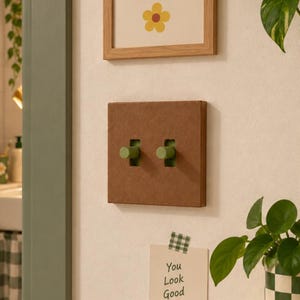 Puede incluir: Una placa de interruptor de luz cuadrada de color marrón con dos interruptores de palanca verdes. La placa está montada en una pared junto a una nota que dice You Look Good Today con un corazón, y el texto NOM NOM OBJECTS está debajo.
