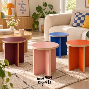May include: Four round side tables with plywood edges and solid-colored surfaces in deep red, light pink, vibrant blue, and bright orange. Each table features a cross-shaped base design. The text "NOM NOM Objects" is printed in black at the bottom.
