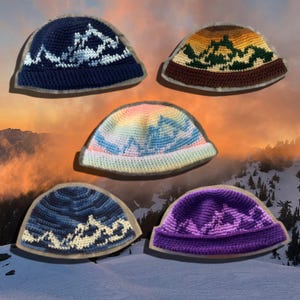 Op de afbeelding: Vijf gehaakte mutsen met bergketenpatronen. De mutsen zijn gekleurd in donkerblauw en lichtblauw, geel en bruin, pastel regenboog, blauw en wit, en paars en donkerpaars. Elke muts heeft een ontwerp met een omgeslagen rand.