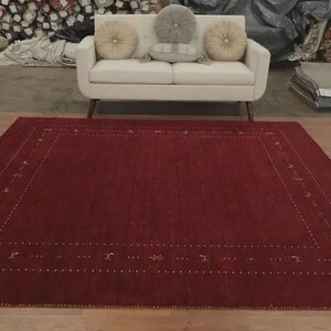 May include: A large, rectangular area rug in a deep, solid red hue. The rug features a decorative border composed of small, white, dotted lines and subtle, spaced-out geometric motifs in muted blue and grey tones, set against the red field.