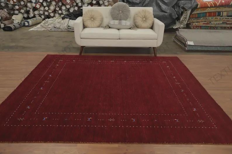 May include: A large, rectangular area rug in a deep, solid red hue. The rug features a decorative border composed of small, white, dotted lines and subtle, spaced-out geometric motifs in muted blue and grey tones, set against the red field.