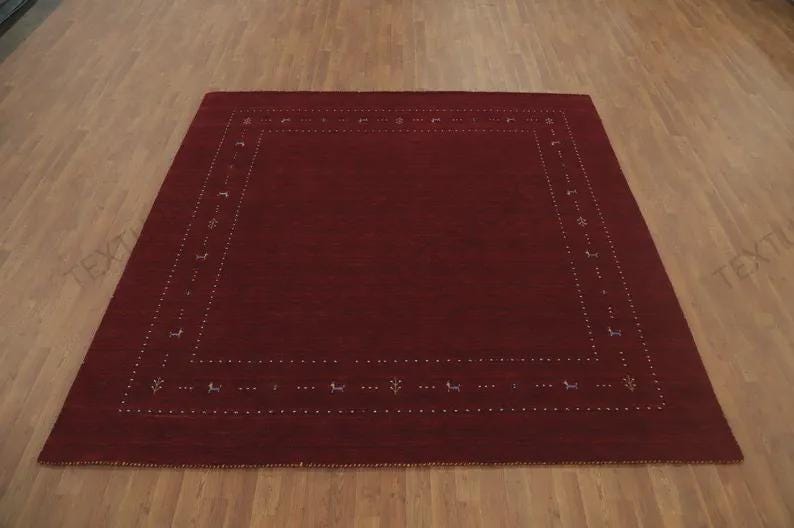 May include: A square area rug in a deep solid red color featuring a decorative border. The border consists of a series of small white dots arranged in a rectangular frame, interspersed with tiny stylized animal and floral motifs in light colors.