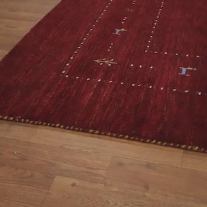 May include: A deep red area rug featuring a rectangular border composed of small white dots. The rug includes small, scattered blue and yellow embroidered motifs of animals and trees within the border. The edges are finished with a thick, braided trim.