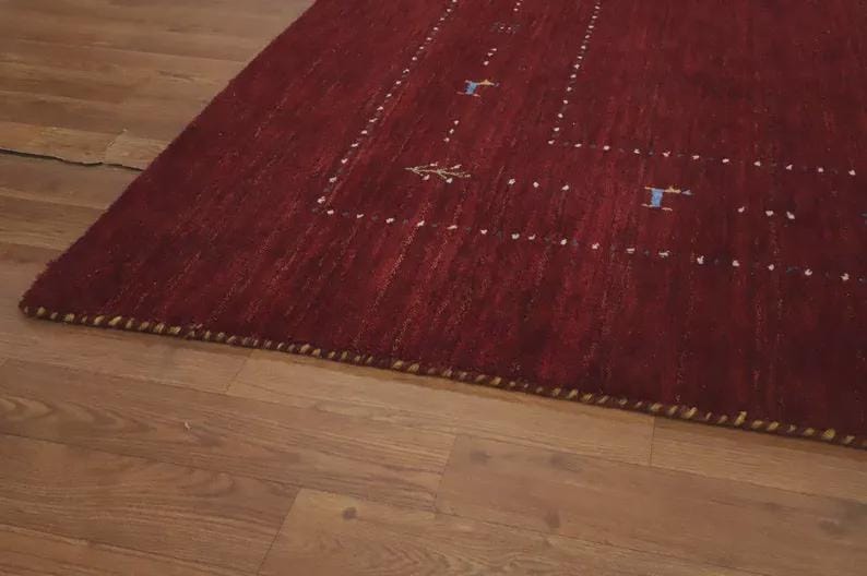 May include: A deep red area rug featuring a rectangular border composed of small white dots. The rug includes small, scattered blue and yellow embroidered motifs of animals and trees within the border. The edges are finished with a thick, braided trim.