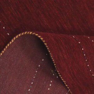 May include: A deep red rug featuring a subtle pattern of small, scattered white and blue geometric accents. The rug is folded over to reveal its underside, showcasing a thick, braided yellow and brown edge binding along the curved fold.