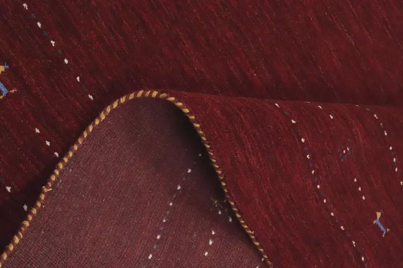 May include: A deep red rug featuring a subtle pattern of small, scattered white and blue geometric accents. The rug is folded over to reveal its underside, showcasing a thick, braided yellow and brown edge binding along the curved fold.