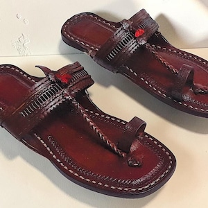 May include: A pair of dark brown leather sandals featuring a toe post, a braided strap, and a wide horizontal band across the midfoot. The edges are finished with white stitching, and a small red accent is visible on the top of each sandal.