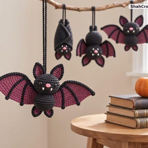 May include: Four crocheted bat ornaments in black and dark purple yarn, featuring small pink noses and white fangs. Three bats hang from a branch, while one hangs in the foreground. A small orange pumpkin sits on a stack of three books nearby.