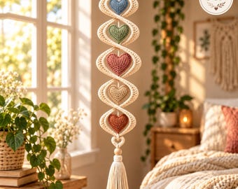 May include: A vertical macrame wall hanging featuring a twisted cream-colored cord structure with five crocheted hearts in blue, green, dusty rose, beige, and terracotta colors, finished with a long cream-colored tassel at the bottom.