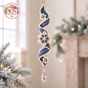May include: A vertical hanging ornament featuring a spiralling blue and cream macrame ribbon adorned with small cream-coloured flowers, a central star-shaped floral motif with a pearl centre, and a clear, faceted teardrop crystal at the bottom.