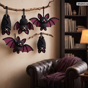 May include: Five crocheted bats in black and dark purple yarn hanging from two wooden branches. The bats feature small white fangs, pink noses, and pointed ears. The text ShahCrochetNest is visible in the upper right corner of the image.