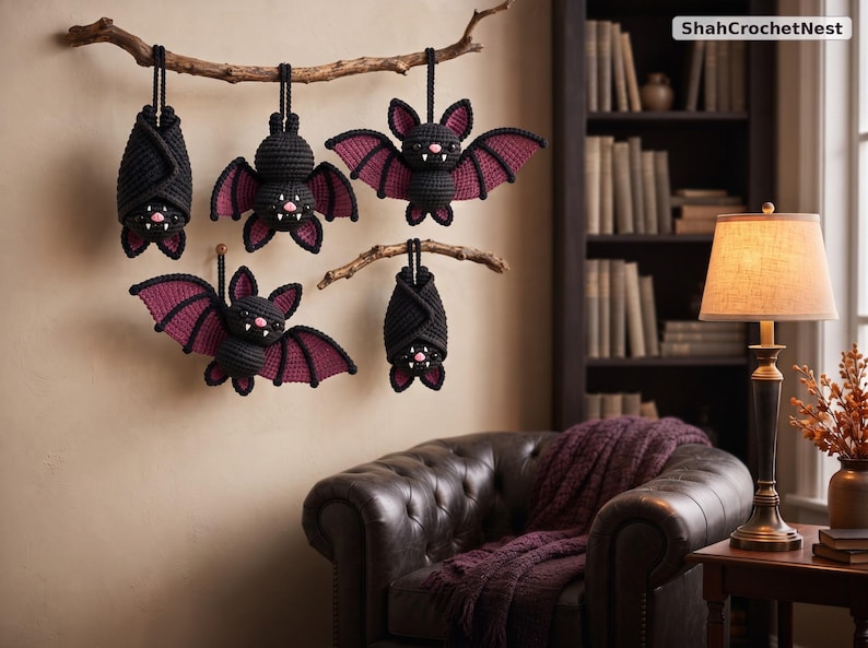 May include: Five crocheted bats in black and dark purple yarn hanging from two wooden branches. The bats feature small white fangs, pink noses, and pointed ears. The text ShahCrochetNest is visible in the upper right corner of the image.