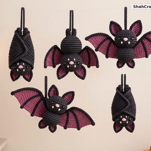 May include: Five crocheted bat ornaments hanging from a string, featuring black bodies with dark purple wing accents, pink noses, and white fangs. Two bats are hanging upside down with wings wrapped around them, while three are displayed with wings spread.