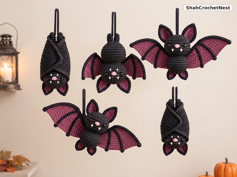 May include: Five crocheted bat ornaments hanging from a string, featuring black bodies with dark purple wing accents, pink noses, and white fangs. Two bats are hanging upside down with wings wrapped around them, while three are displayed with wings spread.