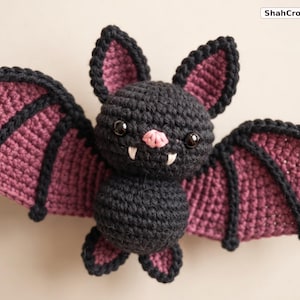 May include: A black and dark purple crocheted bat plush toy with large pointed ears, two white fangs, and a small pink nose. The bat has black plastic eyes and wide, spread wings with black trim. The text ShahCrochetNest is visible in the corner.