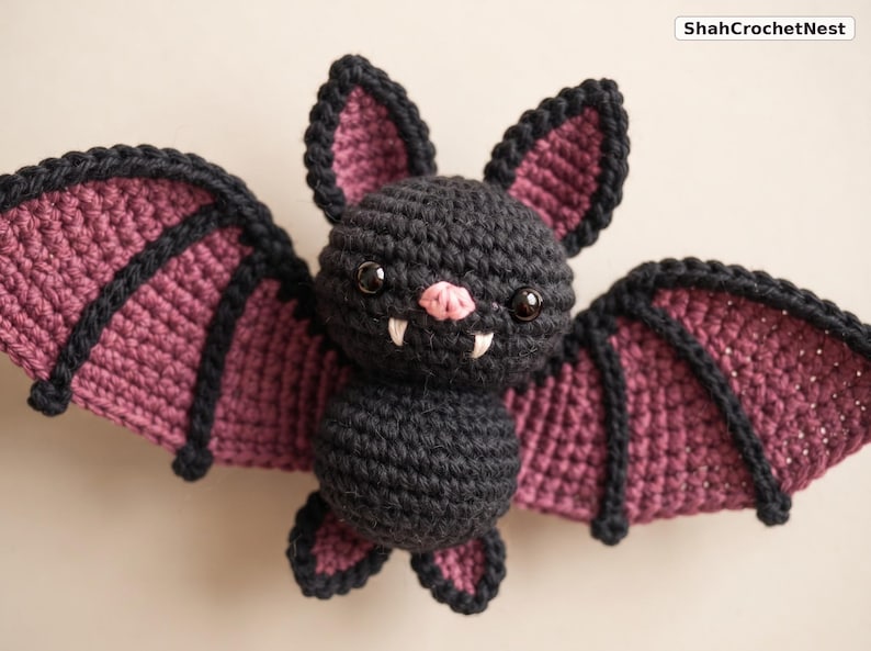 May include: A black and dark purple crocheted bat plush toy with large pointed ears, two white fangs, and a small pink nose. The bat has black plastic eyes and wide, spread wings with black trim. The text ShahCrochetNest is visible in the corner.