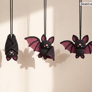 May include: Three crocheted bat ornaments hanging from black cords, labeled ShahCrochetNest. The bats are black with purple inner wings, pink noses, and white fangs. One bat is wrapped in its wings, while the other two have wings extended.