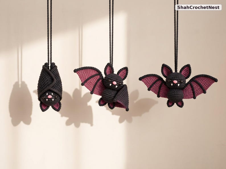 May include: Three crocheted bat ornaments hanging from black cords, labeled ShahCrochetNest. The bats are black with purple inner wings, pink noses, and white fangs. One bat is wrapped in its wings, while the other two have wings extended.