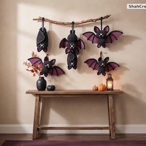 May include: A set of five crocheted bats in black and dark purple yarn hanging from a natural wooden branch. The bats feature small pink noses and white fangs. The text ShahCrochetNest is visible in the upper right corner of the image.