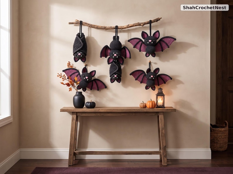 May include: A set of five crocheted bats in black and dark purple yarn hanging from a natural wooden branch. The bats feature small pink noses and white fangs. The text ShahCrochetNest is visible in the upper right corner of the image.