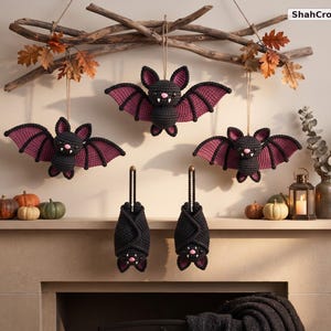 May include: Three crocheted bats with black bodies and purple wing accents hanging from a wooden branch, and two additional crocheted bats hanging vertically below. The bats feature small white fangs and pink noses. Text reads ShahCrochetNest.
