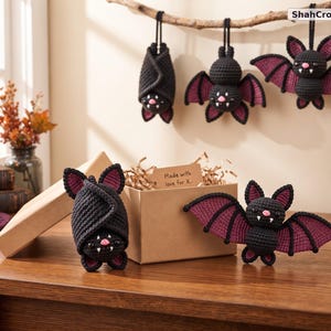 May include: Five crocheted bat plushies in black and dark purple yarn. Three bats hang from a branch, while two sit near a cardboard box with a tag reading Made with love for X. Each bat features small pink noses, white fangs, and pointed ears.