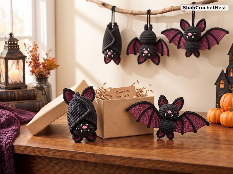 May include: Five crocheted bat plushies in black and dark purple yarn. Three bats hang from a branch, while two sit near a cardboard box with a tag reading Made with love for X. Each bat features small pink noses, white fangs, and pointed ears.