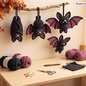 May include: Four crocheted bats in black and dark purple yarn hang from a branch. The bats feature small pink noses and white fangs. Below, on a wooden surface, are balls of black and purple yarn, a crochet hook, scissors, and stitch markers. Text reads ShahCrochetNest.