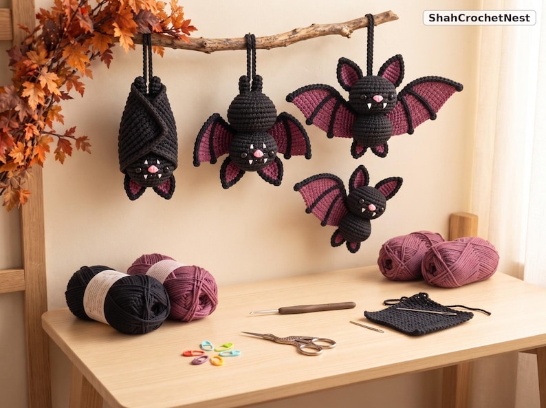 May include: Four crocheted bats in black and dark purple yarn hang from a branch. The bats feature small pink noses and white fangs. Below, on a wooden surface, are balls of black and purple yarn, a crochet hook, scissors, and stitch markers. Text reads ShahCrochetNest.