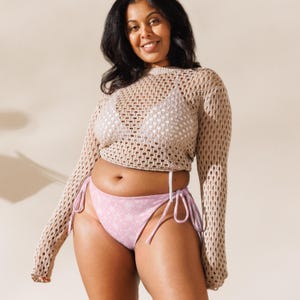 May include: A beige long-sleeve crochet crop top with a wide-knit pattern worn over a white triangle bikini top, paired with light pink bikini bottoms featuring a white floral print and side-tie closures with thin fabric strings.
