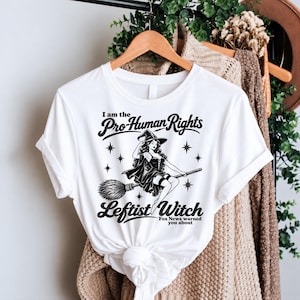 May include: A white t-shirt featuring a black graphic of a witch flying on a broomstick. The text reads: I am the Pro-Human Rights Leftist Witch Fox News warned you about. The design includes small star accents surrounding the central witch figure.