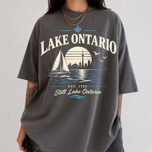 May include: A charcoal gray short-sleeve t-shirt featuring a graphic print of a sailboat on water with a city skyline silhouette, a sun, and birds. The text reads LAKE ONTARIO, EST. 1792, and Still Lake Ontario, with a small blue maple leaf icon.
