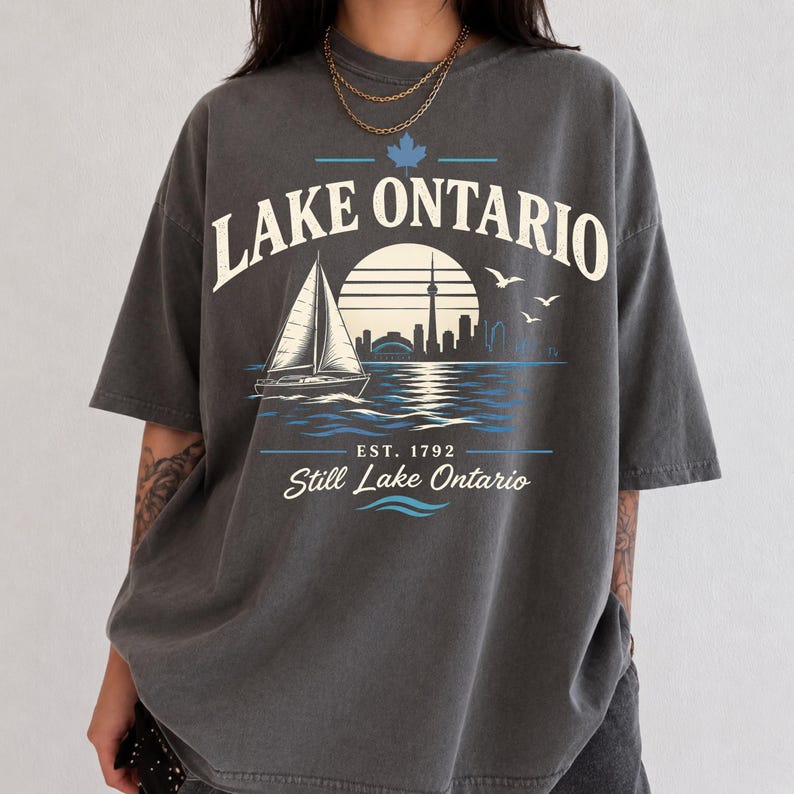 May include: A charcoal gray short-sleeve t-shirt featuring a graphic print of a sailboat on water with a city skyline silhouette, a sun, and birds. The text reads LAKE ONTARIO, EST. 1792, and Still Lake Ontario, with a small blue maple leaf icon.