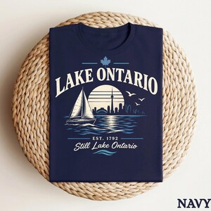 May include: A navy blue t-shirt featuring a graphic of a sailboat on water with a city skyline and sun in the background. The text reads LAKE ONTARIO, EST. 1792, and Still Lake Ontario, accompanied by a maple leaf icon and three flying birds.