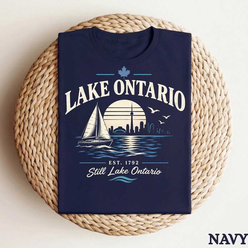 May include: A navy blue t-shirt featuring a graphic of a sailboat on water with a city skyline and sun in the background. The text reads LAKE ONTARIO, EST. 1792, and Still Lake Ontario, accompanied by a maple leaf icon and three flying birds.