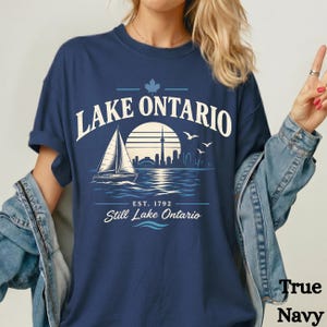 May include: A navy blue t-shirt featuring a white graphic of a sailboat on water, a city skyline, a sun, and birds. The text reads LAKE ONTARIO, EST. 1792, and Still Lake Ontario, with a maple leaf icon above the text. The shirt has a crew neckline.