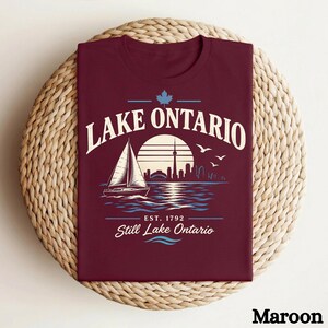 May include: A maroon t-shirt featuring a graphic of a sailboat on water with the Toronto skyline, a maple leaf, and the text "LAKE ONTARIO EST. 1792 Still Lake Ontario" in white and light blue. The design includes a sun and flying birds.