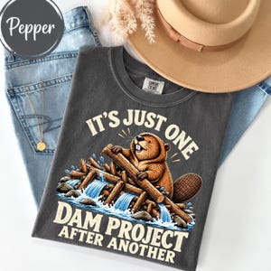 Puede incluir: Una camiseta gris carbón con un gráfico de un castor marrón construyendo una presa de madera con agua azul fluyendo. El texto dice IT'S JUST ONE DAM PROJECT AFTER ANOTHER. La camiseta se presenta con vaqueros azul claro y un sombrero beige.