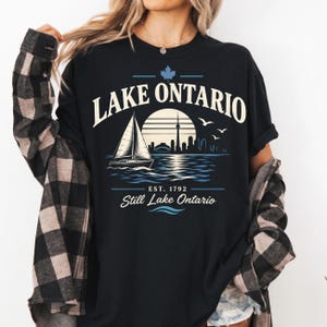May include: A black t-shirt featuring a graphic print of a sailboat on water with a city skyline, a sun, and birds. The text reads LAKE ONTARIO, EST. 1792, and Still Lake Ontario, with a maple leaf icon above the text in a light blue and cream color.