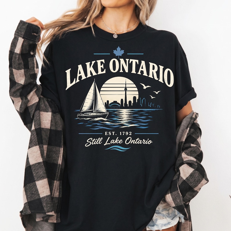 May include: A black t-shirt featuring a graphic print of a sailboat on water with a city skyline, a sun, and birds. The text reads LAKE ONTARIO, EST. 1792, and Still Lake Ontario, with a maple leaf icon above the text in a light blue and cream color.