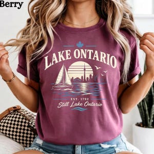 May include: A berry-colored t-shirt featuring a graphic of a sailboat on Lake Ontario, a city skyline, a sun, and birds. The text reads Lake Ontario, Est. 1792, and Still Lake Ontario. The design includes a maple leaf icon above the text.