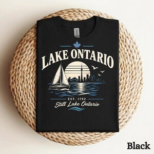 May include: A black t-shirt featuring a graphic of a sailboat on water with a city skyline silhouette, a sun, and birds. The text reads LAKE ONTARIO, EST. 1792, and Still Lake Ontario, with a small maple leaf icon above the text.